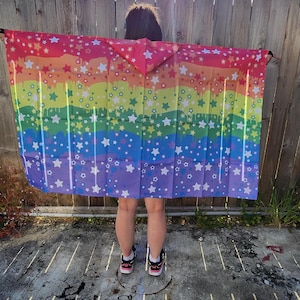 Puede incluir: Una toalla de playa con los colores del arcoíris con un diseño de estrellas y un efecto de pintura goteando. La toalla se sostiene contra una valla de madera y un cielo soleado. Los colores de la toalla incluyen rojo, naranja, amarillo, verde, azul y morado.