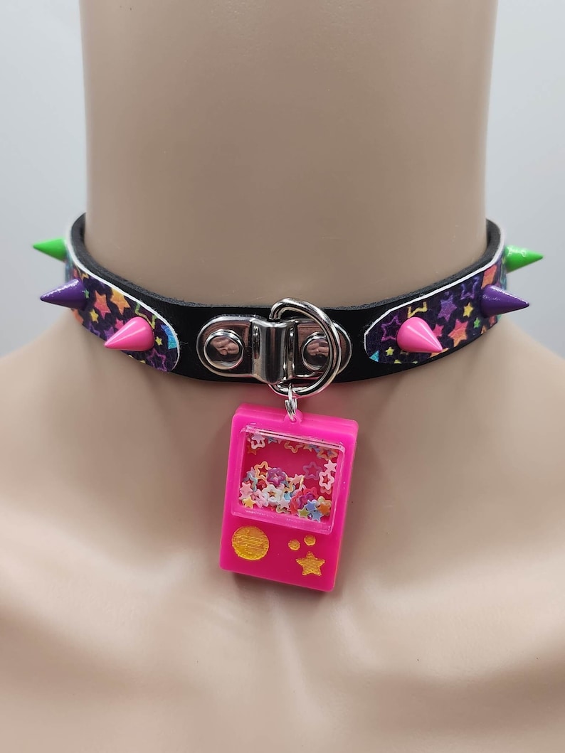 Pu&ograve; includere: Un collare choker nero con dettagli argentati e una stampa di stelle colorate. Il choker ha punte verdi e rosa. Un ciondolo rosa con una finestra trasparente piena di stelle colorate &egrave; appeso al collare.