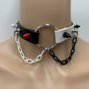 May include: A black and white leather choker with silver rings and spikes. The choker has two chains attached, one white and one black.