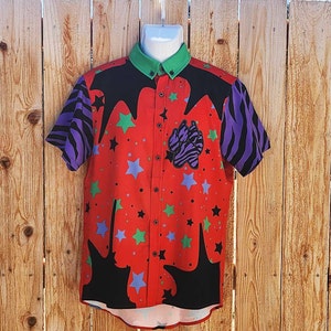 May include: A short-sleeved button-down shirt with a vibrant red base, adorned with a pattern of blue and green stars, black drips, and a zebra-striped paw print. The sleeves feature purple and black zebra stripes, and the collar is green.