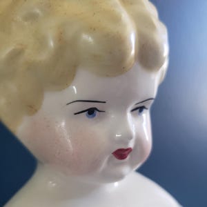 May include: A porcelain doll head featuring molded blonde hair with a textured finish, painted blue eyes, defined black eyebrows, rosy pink cheeks, and bright red lips. The facial features are smooth and glossy, capturing a detailed expression.