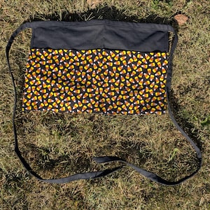 May include: A black waist apron with a candy corn pattern. The apron features a black top section and a lower section covered in a repeating pattern of yellow, orange, and white candy corn on a black background. Long black ties extend from the sides.