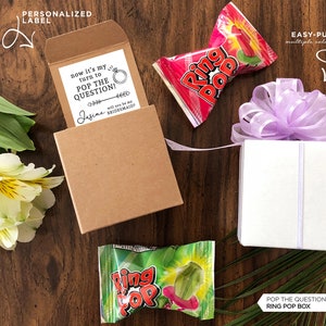 May include: A brown box with a white card inside that says "now it's my turn to POP THE QUESTION!" with a ring and arrow graphic. The card is addressed to "Jasmine" and says "Will you be my bridesmaid?"  The box is sitting on a wooden surface with a Ring Pop sweet and a white box with a purple bow on top.