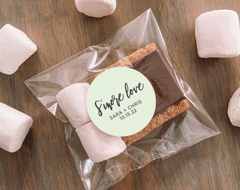 Puede incluir: Una bolsa de plástico transparente llena de dos malvaviscos blancos y un s'more. El s'more está hecho con una galleta graham, chocolate y un malvavisco. Una pegatina circular verde con el texto "S'more love SARA + CHRIS 10.15.22" está pegada a la bolsa.