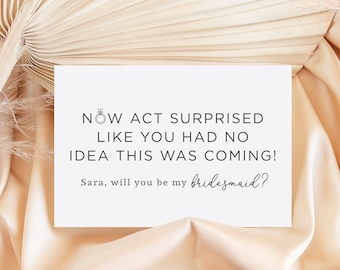 Op de afbeelding: Een witte kaart met de tekst "NOW ACT SURPRISED LIKE YOU HAD NO IDEA THIS WAS COMING! Sara, will you be my bridesmaid?" erop gedrukt. De kaart ligt op een beige stoffen achtergrond.