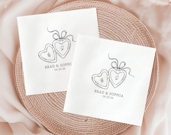 May include: Two white square napkins with a heart-shaped locket design and the names "Brad & Sophia" and the date "06.20.26" printed in black. The napkins are placed on a woven, circular placemat.