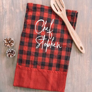 May include: Red and black buffalo plaid kitchen towel with a white embroidered design that says "Chef Stephen". A wooden spoon is lying on top of the towel.