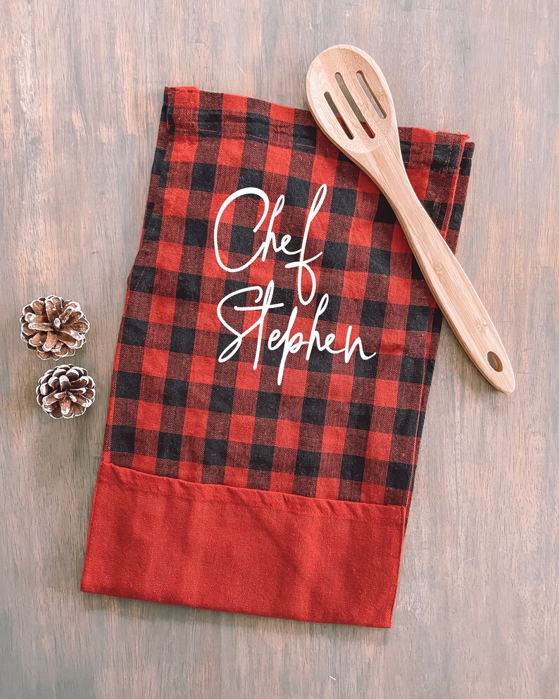 May include: Red and black buffalo plaid kitchen towel with a white embroidered design that says "Chef Stephen". A wooden spoon is lying on top of the towel.