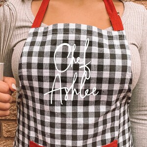 May include: Black and white checkered apron with red straps and a red pocket. The apron has the text "Chef Ashlee" written in white cursive.