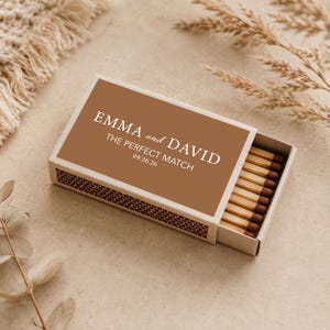 Puede incluir: Una caja de cerillas con tapa marrón y caja blanca. La tapa tiene el texto blanco "EMMA and DAVID THE PERFECT MATCH 09.26.26". La caja está parcialmente abierta, revelando cerillas de madera con puntas marrones.