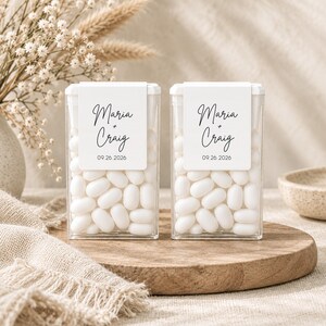 Op de afbeelding: Twee doorzichtige, rechthoekige plastic bakjes gevuld met witte, ovale snoepjes. Elk bakje is voorzien van een wit label met de namen Maria + Craig en de datum 26-09-2026 in een zwarte combinatie van script- en schreefloze lettertypes.