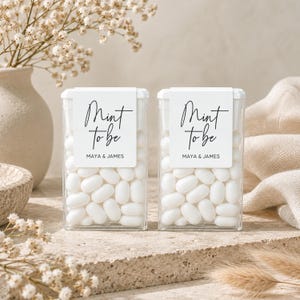May include: Two clear rectangular plastic containers filled with white oval mints. Each container features a white label with the black text Mint to be and the names Maya & James printed in a clean, modern sans-serif font below the script text.