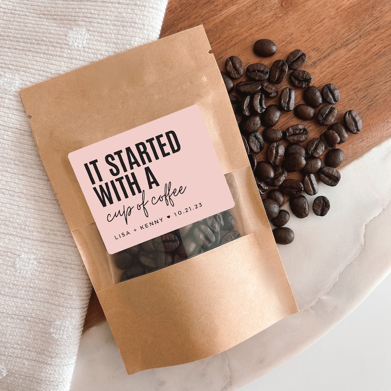 K&ouml;nnte beinhalten: Ein brauner Papierbeutel mit einem rosa Etikett, auf dem "It started with a cup of coffee" und "Lisa + Kenny 10.21.23" steht. Der Beutel ist mit Kaffeebohnen gef&uuml;llt.