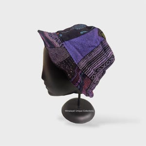 Op de afbeelding: Een patchwork emmerhoed in tinten paars, zwart en blauw. De hoed wordt getoond op een zwarte mannequinhoofd. De hoed heeft verschillende stofpatronen, waaronder geometrische en gestreepte ontwerpen. De rand is iets omhoog geklapt.