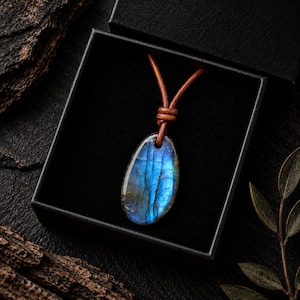 May include: A polished, teardrop-shaped labradorite pendant featuring vibrant blue iridescence and dark inclusions. The stone is suspended from a brown leather cord that is knotted at the top, presented inside a square black gift box.