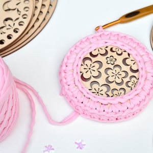 May include: Pink yarn is wrapped around a wooden circle with a floral design. The yarn is being crocheted around the circle to create a round, pink crocheted coaster.