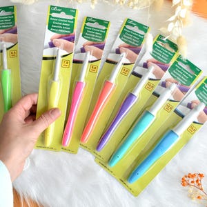 May include: A collection of Clover Amour crochet hooks in various sizes and vibrant colors, including green, yellow, pink, orange, purple, and blue. Each hook is packaged on a yellow and green card with the hook size clearly marked in millimeters.