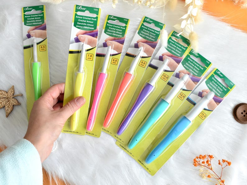 May include: A collection of Clover Amour crochet hooks in various sizes and vibrant colors, including green, yellow, pink, orange, purple, and blue. Each hook is packaged on a yellow and green card with the hook size clearly marked in millimeters.