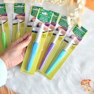 May include: A collection of Clover Amour crochet hooks in various sizes, displayed on their original packaging. The hooks have colorful handles in shades of green, yellow, blue, purple, and teal. The packaging includes the text "Amour Crochet Hook".