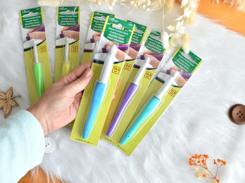 May include: A collection of Clover Amour crochet hooks in various sizes, displayed on their original packaging. The hooks have colorful handles in shades of green, yellow, blue, purple, and teal. The packaging includes the text "Amour Crochet Hook".