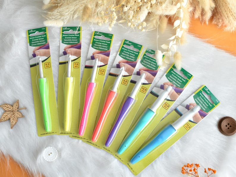 May include: A collection of Clover Amour crochet hooks in various sizes, each with a colorful, ergonomic handle. The hooks range from 6.5 mm to 15.0 mm, displayed on a white surface with decorative elements.