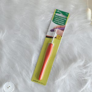 May include: An orange and silver crochet hook, labeled "Amour Crochet Hook" with a 3.0 mm size indicator, is displayed on a green and white package. The hook is set against a white, fluffy background.