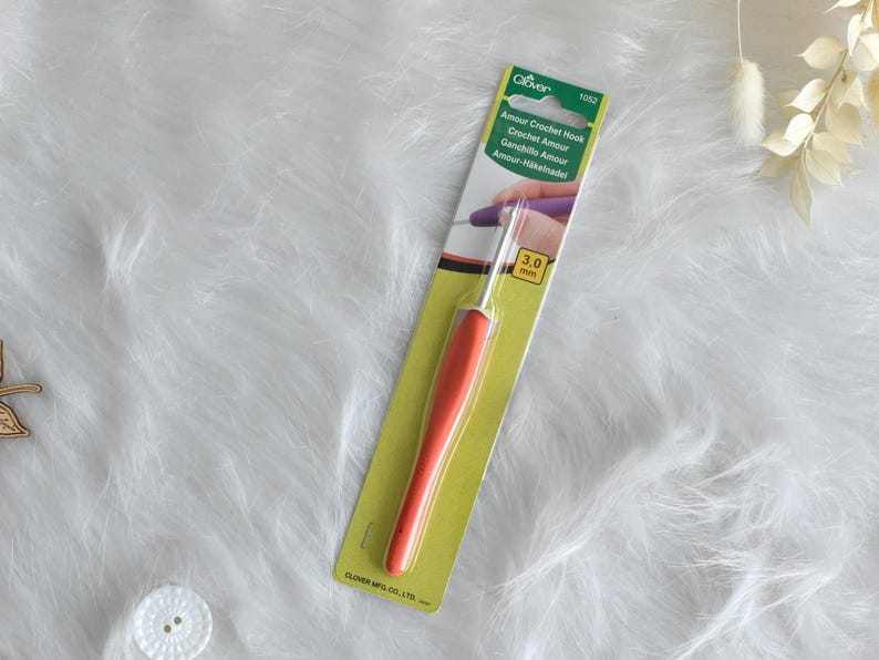 May include: An orange and silver crochet hook, labeled "Amour Crochet Hook" with a 3.0 mm size indicator, is displayed on a green and white package. The hook is set against a white, fluffy background.