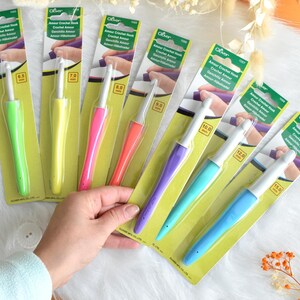 May include: A collection of Clover Amour crochet hooks in various sizes and colors. The hooks have ergonomic handles in shades of green, yellow, pink, orange, purple, teal, and blue. Each hook is packaged on a yellow and green card with product information.