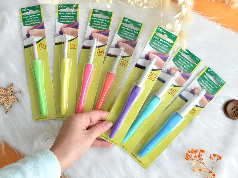 May include: A collection of Clover Amour crochet hooks in various sizes and colors. The hooks have ergonomic handles in shades of green, yellow, pink, orange, purple, teal, and blue. Each hook is packaged on a yellow and green card with product information.