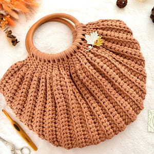 May include: A handmade, peach-colored crochet bag with wooden handles. The bag features a leaf-shaped embellishment and a textured, ribbed design. A ball of yarn, crochet hook, and scissors are also visible.