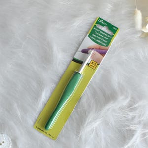 May include: A green and silver crochet hook on a yellow and white package. The package has the text "Amour Crochet Hook" and "5.5 mm". The hook is designed for crochet projects.