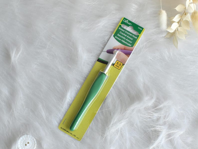 May include: A green and silver crochet hook on a yellow and white package. The package has the text "Amour Crochet Hook" and "5.5 mm". The hook is designed for crochet projects.