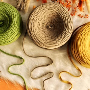 May include: Three skeins of yarn in shades of green, tan, and yellow are displayed. The yarn is arranged on a white, fluffy surface, with a wooden leaf and buttons scattered around. The skeins are unwound, with the yarn extending from each.