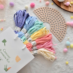 Puede incluir: Una bolsa de papel blanca con la etiqueta "Crochet Rainbow Kit" contiene madejas de hilo colorido en tonos morado, azul, verde, amarillo, rosa y crema. También se ve una herramienta de madera en forma de abanico y pompones decorativos.