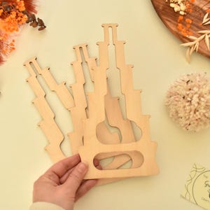 May include: A stack of unfinished, light-colored wooden craft pieces with a unique shape, held by a hand. The pieces have a cutout design. The image also includes dried flowers, a pom-pom, and a business card.