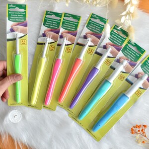 May include: A collection of Clover Amour crochet hooks in various sizes, each with a colorful, ergonomic handle. The hooks range from 6.5 mm to 15.0 mm, displayed on a white, fluffy surface. The packaging is green and yellow.