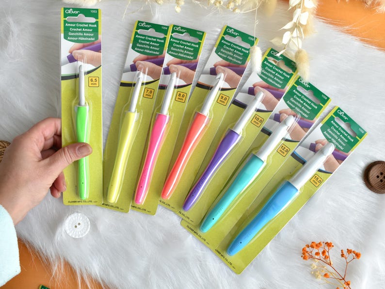 May include: A collection of Clover Amour crochet hooks in various sizes, each with a colorful, ergonomic handle. The hooks range from 6.5 mm to 15.0 mm, displayed on a white, fluffy surface. The packaging is green and yellow.
