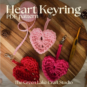 May include: A wooden cutting board displays three handmade heart keyrings in pink and red, alongside a ball of pink yarn and a crochet hook. The keyrings are attached to silver clasps. Text on the image reads "Heart Keyring PDF pattern" and "The Green Lake Craft Studio."