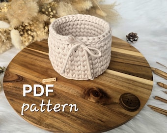 May include: A round, beige crochet basket made from thick cord, featuring a decorative tie detail at the front. The image includes the text PDF pattern, indicating this is a digital guide for creating the item shown in the photograph.