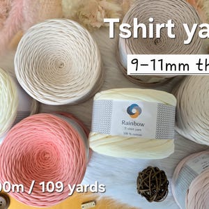 Può includere: Un assortimento di filato per t-shirt nei colori crema, rosa e beige. L'immagine mostra il testo "Tshirt yarn", "9-11mm thick" e "100m / 109 yards". Un'etichetta recita "Rainbow T-shirt yarn 100% cotton". Lo spessore è di 9-11mm.