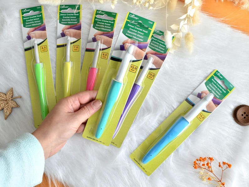May include: A collection of Clover Amour crochet hooks in various sizes and colors, including green, yellow, pink, blue, and purple. Each hook is packaged on a yellow and green card with the hook size in millimeters.