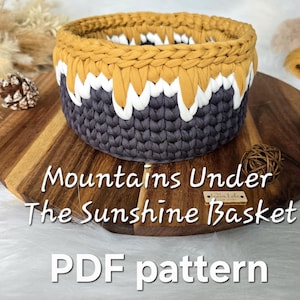 May include: A handmade crochet basket with a unique design. The basket features a color-blocked pattern with mustard yellow, white, and gray sections. The text "Mountains Under The Sunshine Basket" and "PDF pattern" are visible.