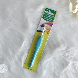 May include: A blue crochet hook with a 4.5 mm size, on a green and yellow card. The card has the text "Amour Crochet Hook" in multiple languages. The hook is lying on a white, fluffy surface.