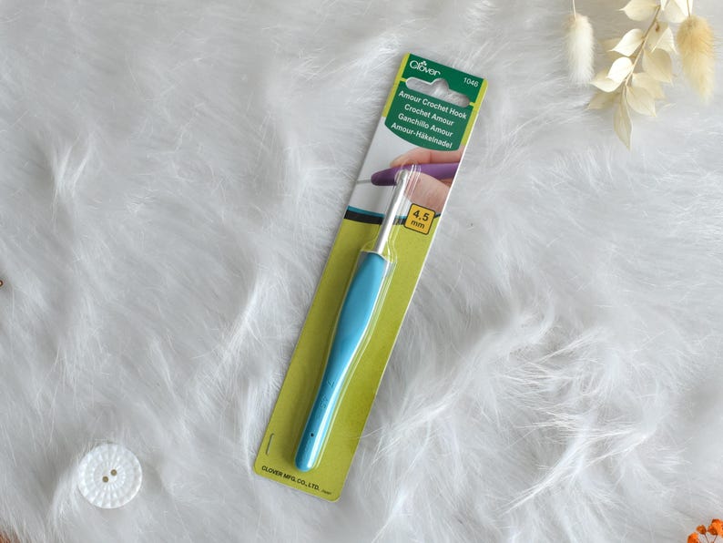 May include: A blue crochet hook with a 4.5 mm size, on a green and yellow card. The card has the text "Amour Crochet Hook" in multiple languages. The hook is lying on a white, fluffy surface.
