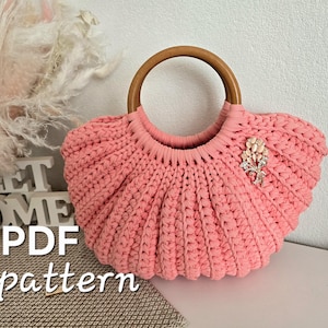 May include: A pink crocheted handbag featuring a rounded wooden handle and a decorative floral brooch with clear crystals. The bag has a textured, ribbed knit pattern and a semi-circular shape. The text "PDF pattern" is visible in the lower left.