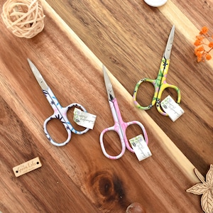 May include: Three pairs of decorative scissors with floral patterns in blue, pink, and yellow, arranged on a wooden surface. The scissors have small tags attached. Other decorative items, including wooden buttons and woven balls, are also present.