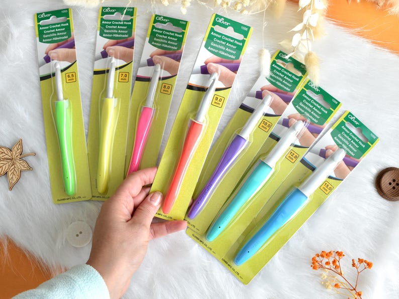 May include: A collection of Clover Amour crochet hooks in various sizes, displayed on their original packaging. The hooks have colorful handles in shades of green, yellow, pink, orange, purple, and blue. The packaging includes the hook size in millimeters.