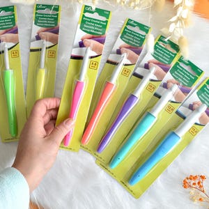 May include: A collection of Clover Amour crochet hooks in various sizes, each with a colorful, ergonomic handle. The hooks range from 6.5 mm to 15.0 mm, displayed on their original packaging. The image shows a hand holding one of the hooks.