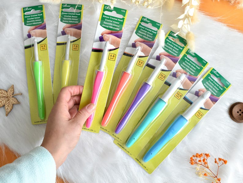 May include: A collection of Clover Amour crochet hooks in various sizes, each with a colorful, ergonomic handle. The hooks range from 6.5 mm to 15.0 mm, displayed on their original packaging. The image shows a hand holding one of the hooks.