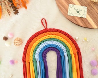 Puede incluir: Un colgante de pared de arcoíris hecho a mano en colores vibrantes de rojo, naranja, amarillo, verde, azul y morado. El arcoíris está hecho de tela tejida y tiene un lazo para colgar. Pompones decorativos y un círculo de madera en el fondo.